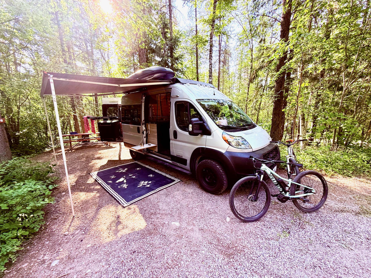Luna parked at a forest campsite with the awning out, the outdoor mat down and a mountain bike leaning beside the van.