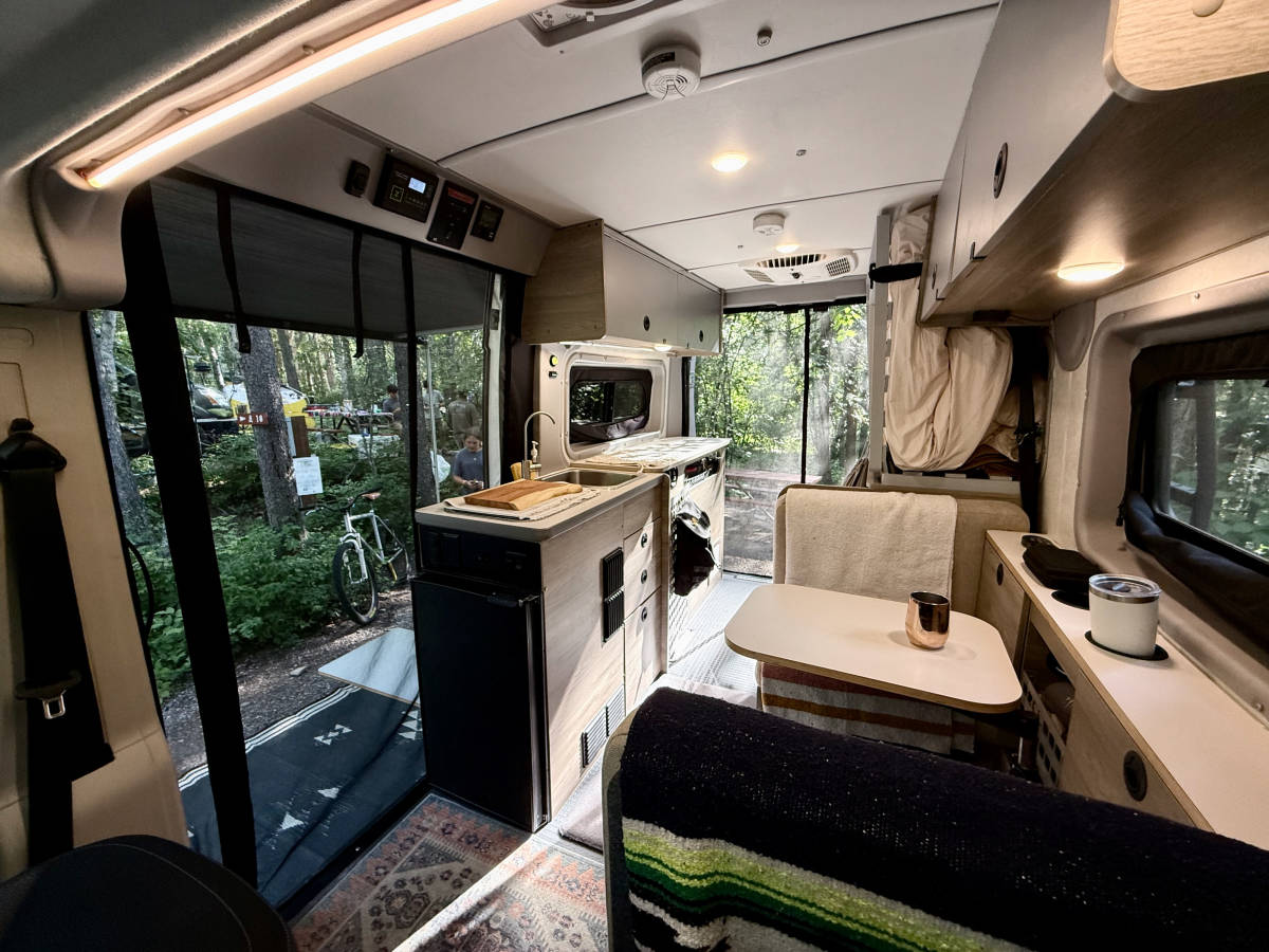 The living space seen from the cab: the sliding door open on the left with a bike outside, the fridge and galley in the middle, the dinette table on the right.