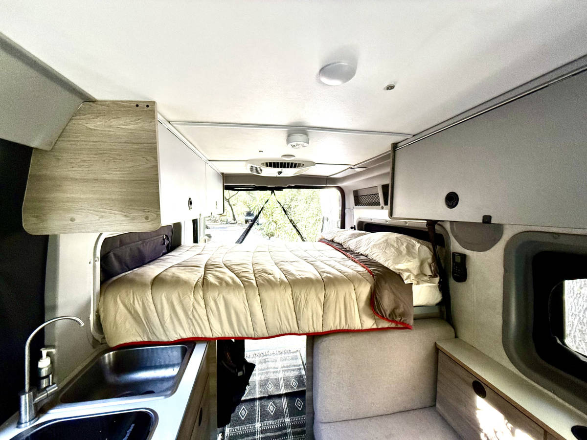 The queen bed folded down and made up, with the rear doors open to the trees behind it.