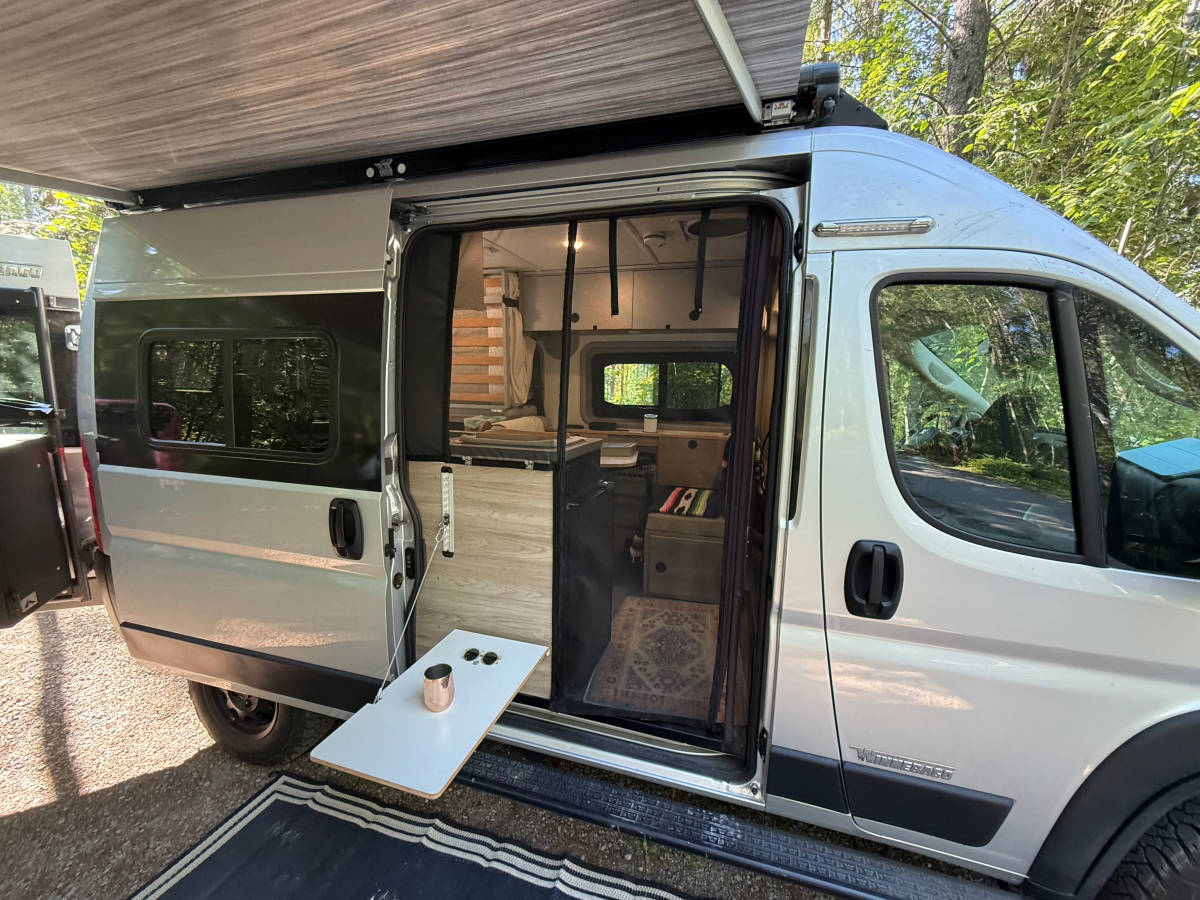 The sliding door open with the bug screen drawn across it, a flip-out table at the sill, the awning overhead and the outdoor mat on the ground.
